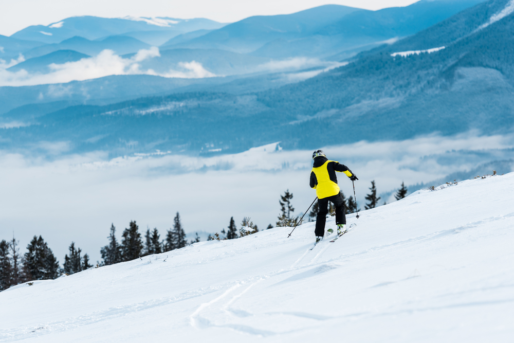 A skier wearing a bright yellow jacket skis downhill on a snowy mountain slope with pine trees, with misty hills and blue mountains in the background.