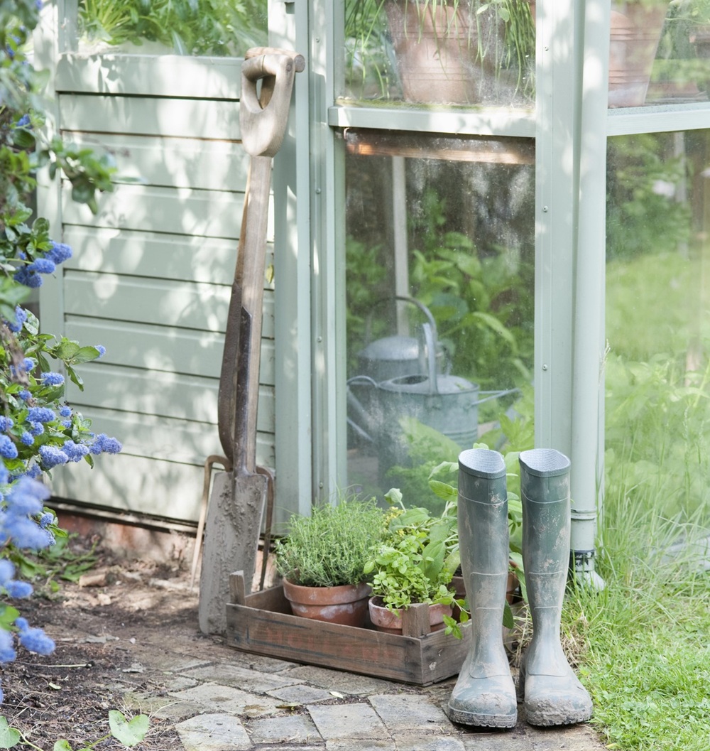 A pair of muddy green wellies, a metal watering can, a garden spade, and a wooden crate of potted herbs sit beside a pale green glasshouse surrounded by lush plants and flowers.