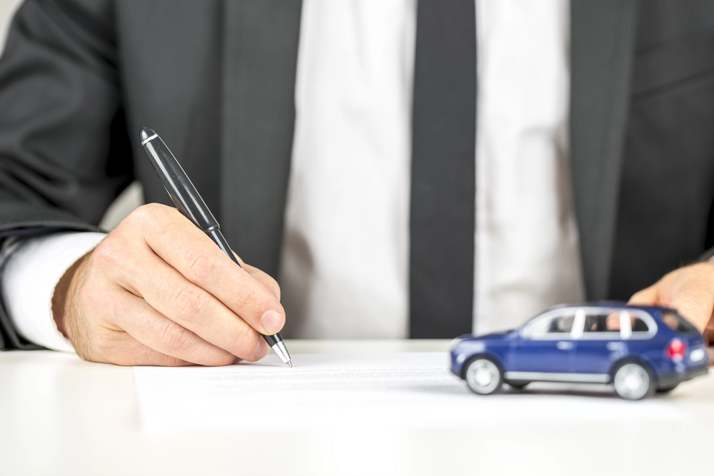 A person in a suit signs a document at a desk, with a small blue toy car placed nearby, symbolising a car purchase or insurance agreement.