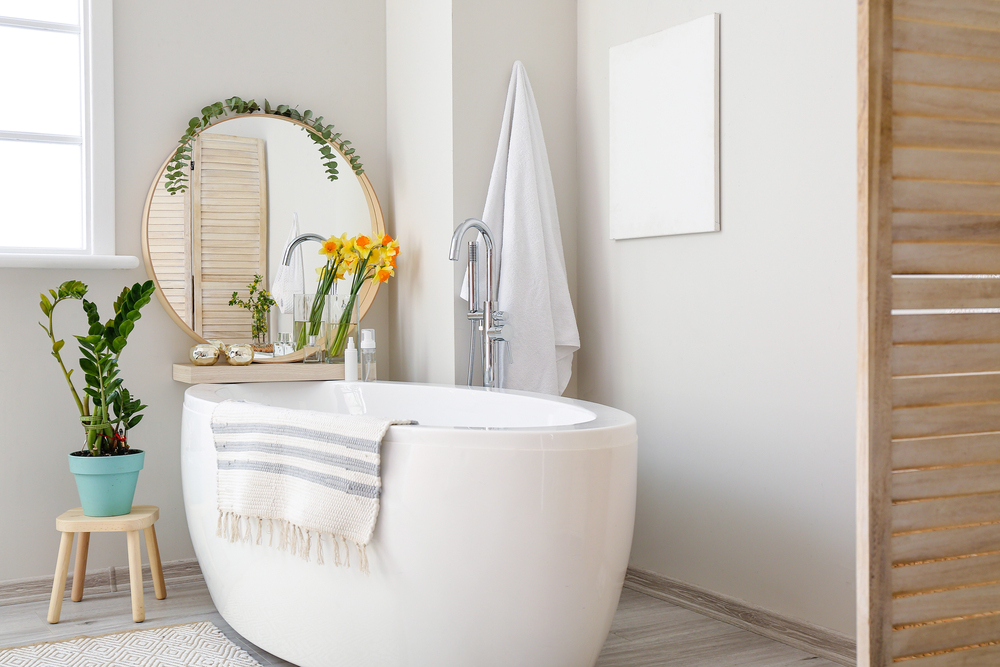 A modern bathroom with a white freestanding bath, a striped towel draped over the edge, a round mirror, green plants, yellow flowers in a vase, and a white towel hanging on the wall. Neutral colours and soft lighting.