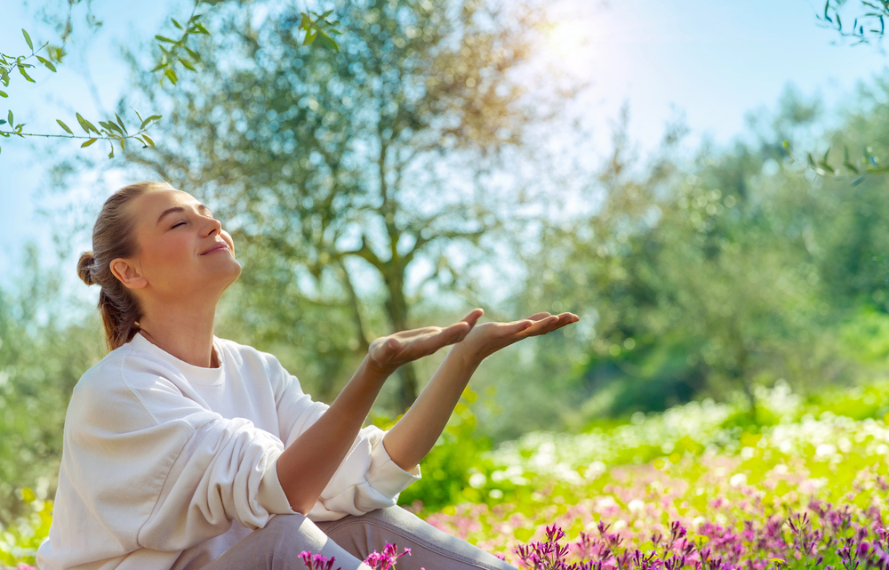 A woman sits outdoors in a sunlit garden with colourful flowers, smiling peacefully with her eyes closed and hands raised, enjoying the nature and sunlight. Trees and greenery surround her in the background.