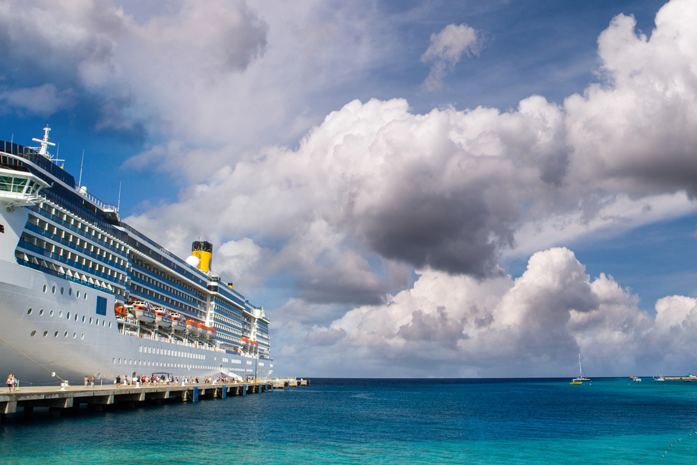 A large cruise ship docked at a pier under a partly cloudy sky, with turquoise blue sea water and a few small boats in the distance.