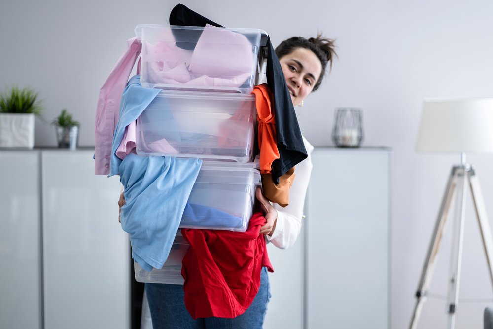 A person smiles while holding stacked clear boxes overflowing with colourful clothes in a bright, modern room with white cupboards, plants, and a tripod lamp in the background.