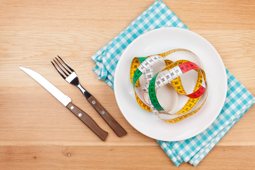 A white plate with a coiled, colourful measuring tape on it sits on a blue checked serviette. A knife and fork are placed to the left of the plate on a wooden table.