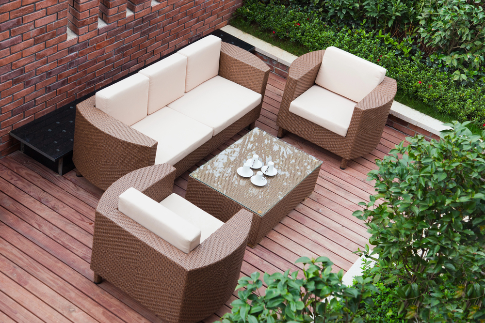 Outdoor patio with a wicker sofa, two armchairs, and a glass-topped coffee table holding cups and saucers, set on a wooden decking surrounded by green bushes and a red brick wall.