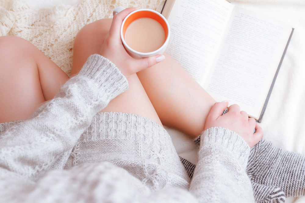A person wearing a cosy light grey jumper sits with crossed legs, holding a cup of coffee or tea in one hand and an open book in the other, creating a relaxed, comfortable atmosphere.