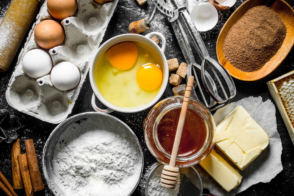 A baking scene with eggs, flour, honey, sugar, cocoa powder, butter, cinnamon sticks, a rolling pin, vintage egg beaters, and biscuit cutters arranged on a dark surface.
