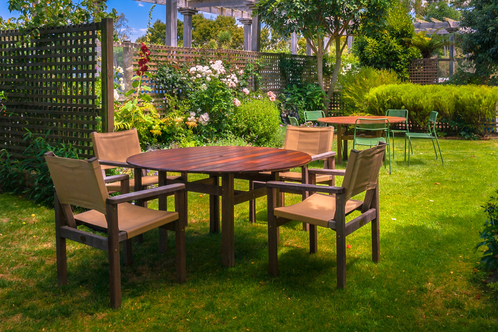 A round wooden table with four beige chairs sits on green grass in a garden, surrounded by flowers, bushes, and trellis fences. Another table and chairs are seen in the background under trees.