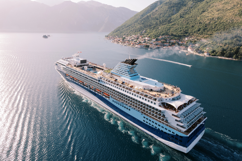 A large white and blue cruise ship sails through calm blue water near a coastal village and green mountains under a clear sky. People are visible on the decks enjoying the view.