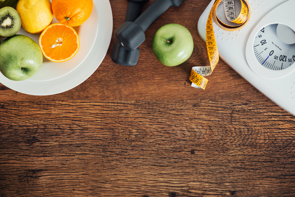 A plate of fresh fruit, two green apples, a pair of black dumbbells, a yellow measuring tape, and a white bathroom scale sit on a wooden surface, symbolising healthy living and fitness.