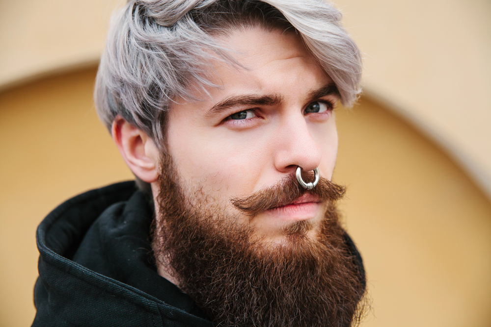A young man with silver hair, a thick brown beard, a curled moustache, and a septum piercing looks at the camera whilst wearing a black hoodie. The background is blurred and yellowish.
