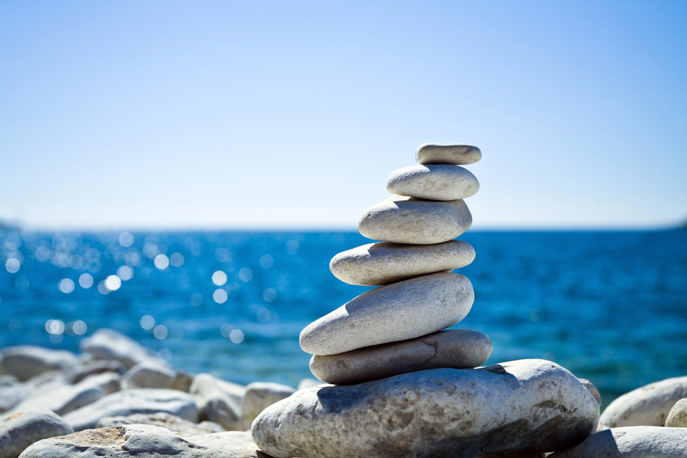 A stack of smooth white stones balanced on a larger rock with a blue, sparkling sea and clear sky in the background. The scene evokes calm and tranquillity.