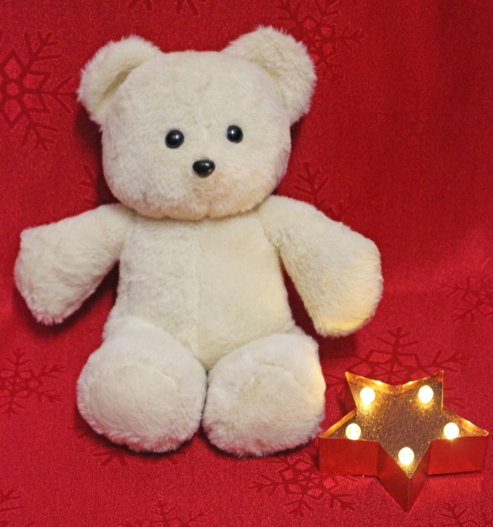 A cream-coloured teddy bear sits on a red surface with snowflake patterns, next to a lit star-shaped decoration with small yellow lights.