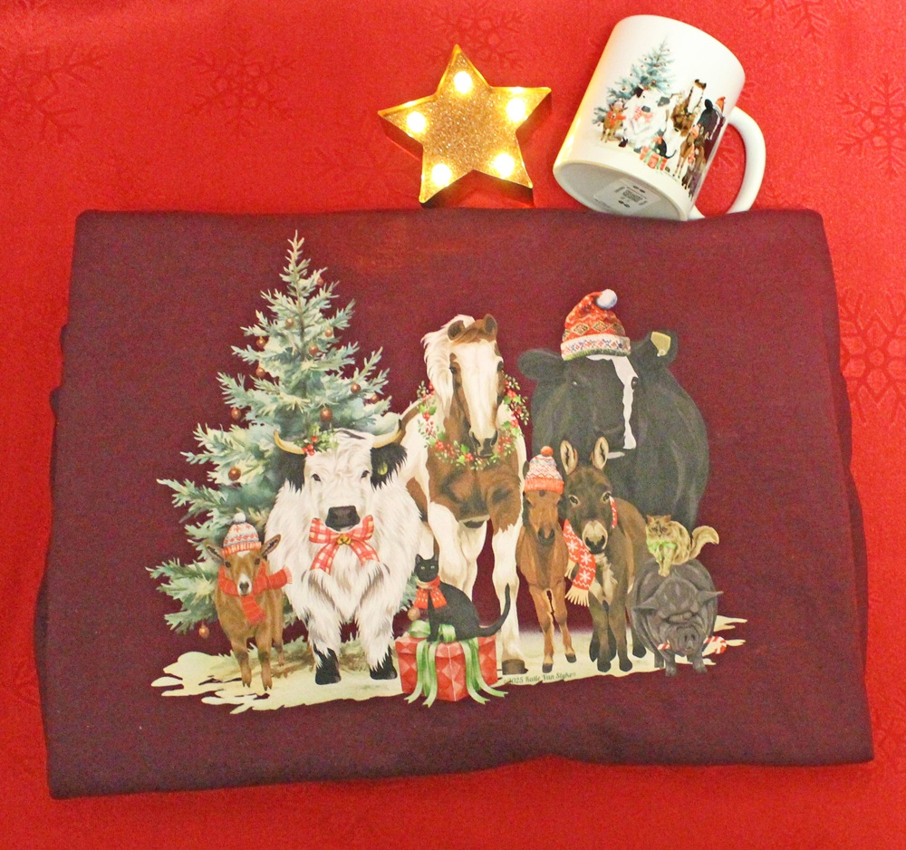 A burgundy shirt with a festive print of farm animals in holiday attire in front of a Christmas tree, with a matching mug and a lit star ornament on a red background.
