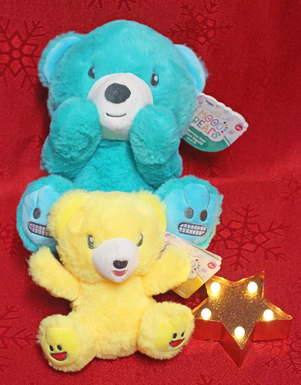 Two plush bears, one blue and one yellow, are stacked on a red background with snowflake patterns. The yellow bear is in front, and a small lit star-shaped decoration is beside them. Both bears have happy facial expressions.