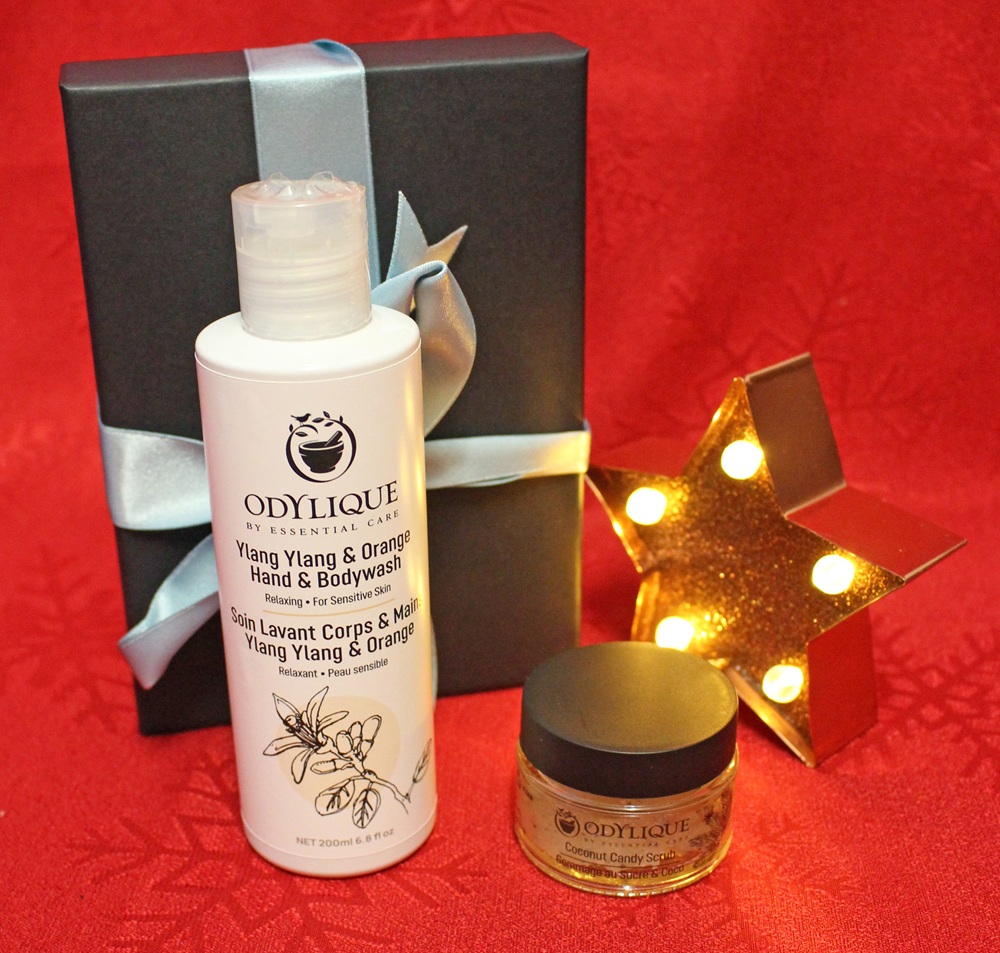 A bottle of Odylique Ylang Ylang & Orange Hand & Body Wash, a small jar of Coconut Candy Scrub, a black gift box with a silver ribbon, and a star-shaped light are arranged on a red background.