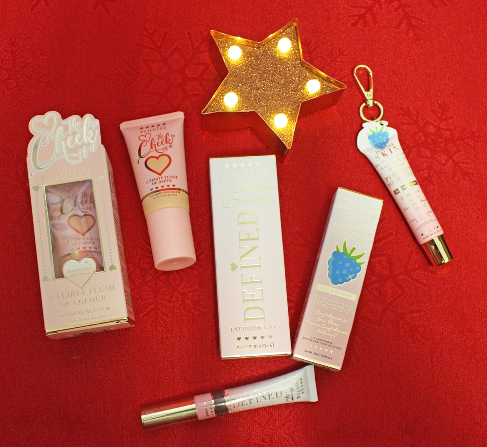Cosmetic products, including blusher tubes and lip gloss, are arranged on a red patterned surface with a lit gold star decoration. Packaging is colourful and decorative, with some items featuring hearts and fruit illustrations.