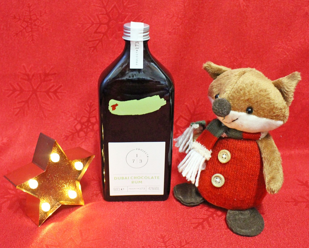 A bottle of Dubai chocolate rum stands on a red patterned surface next to a small plush fox in a red coat and scarf, and a lit star-shaped lamp.