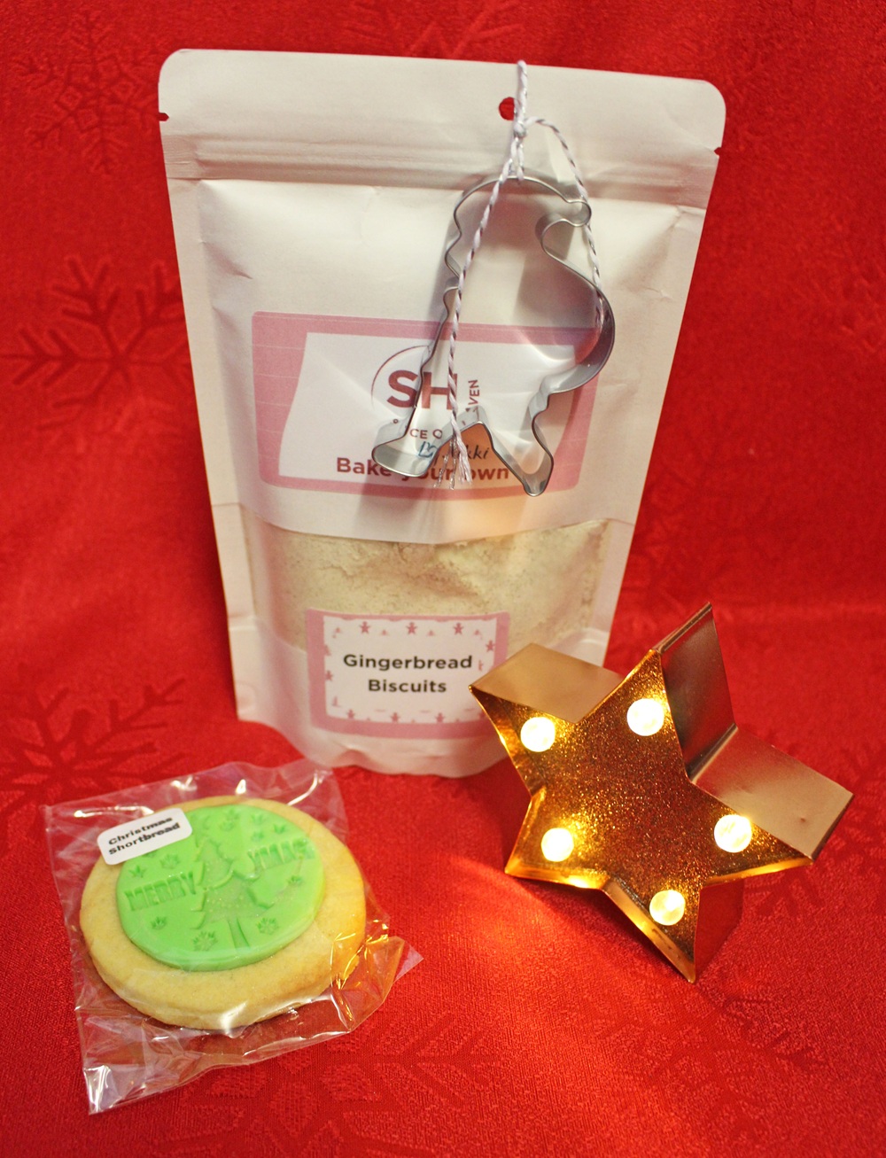 A gingerbread biscuit baking kit with a reindeer biscuit cutter, a packaged decorated biscuit, and a lit star-shaped ornament, all arranged on a red festive background.