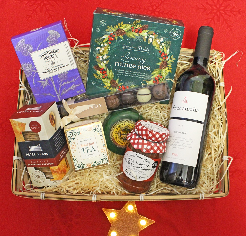 A festive gift hamper on red cloth includes mince pies, shortbread, crispbread, tea, chutney, cheese, truffles, and a bottle of red wine, all arranged neatly on straw with a glowing star-shaped light beneath.