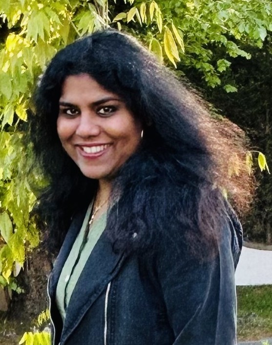 A woman with long, dark, wavy hair smiles at the camera while standing outdoors in front of green foliage. She is wearing a dark jacket over a green top.