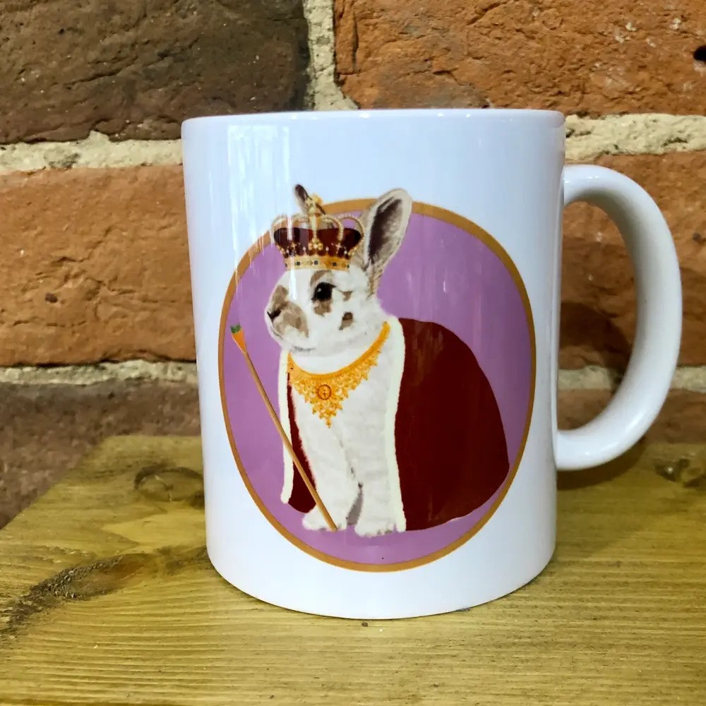 A white mug featuring an illustration of a rabbit dressed as a royal monarch, wearing a crown, cape, and necklace, with a sceptre, set against a purple circular background.