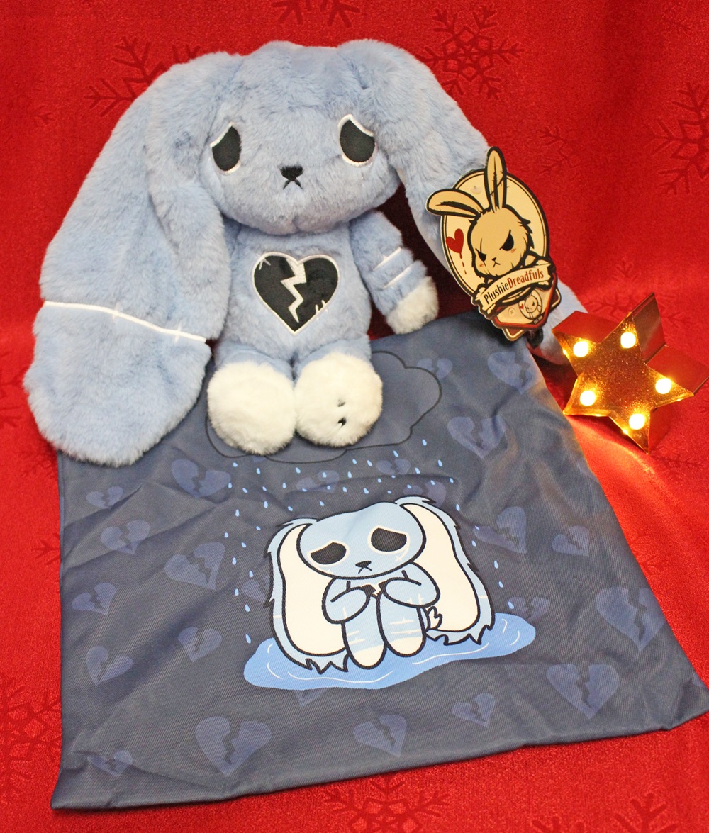 A plush blue bunny with a broken heart on its chest sits on a matching blanket featuring a sad bunny in the rain. A star-shaped light and a cardboard bunny sign are next to the plush, all set against a red background.