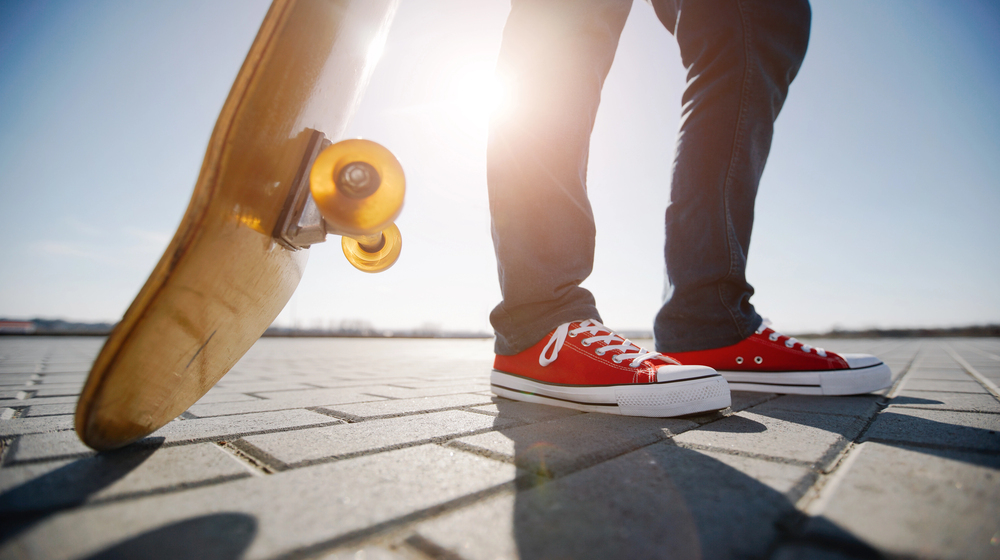 Person wearing red trainers standing on pavement with one foot on a skateboard, sunlight shining in the background, creating a bright and energetic atmosphere.
