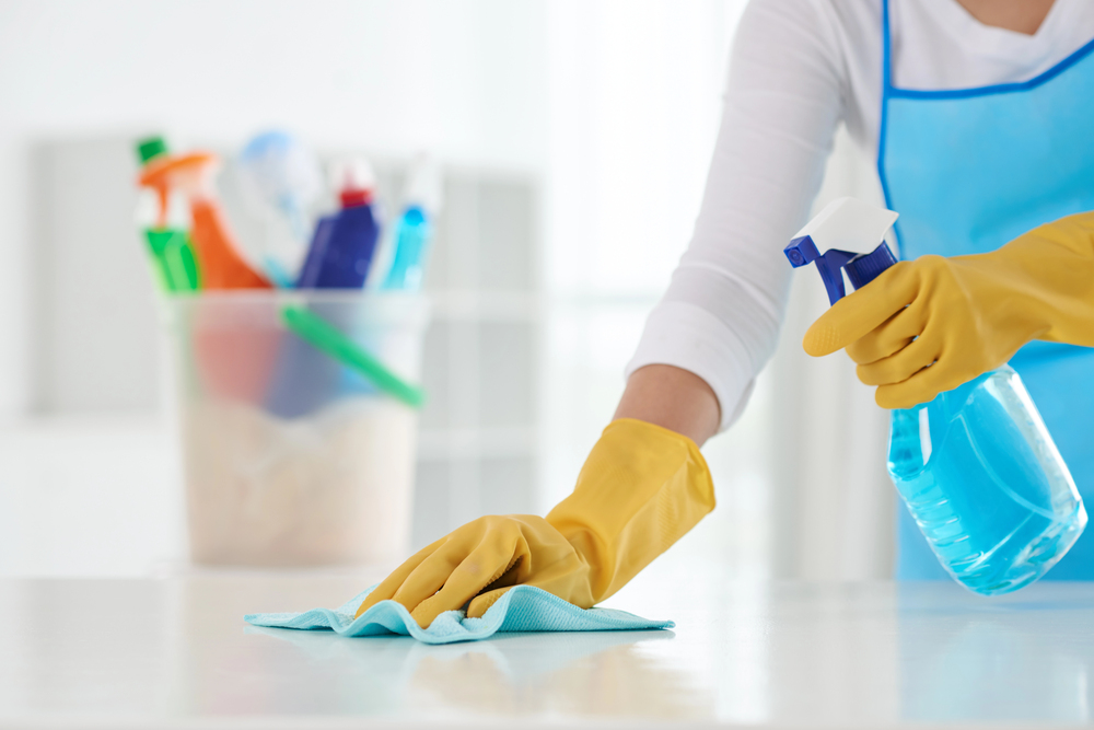 A person wearing yellow rubber gloves cleans a white surface with a blue cloth and holds a spray bottle of cleaning solution. In the background, a bucket filled with various cleaning products is visible.