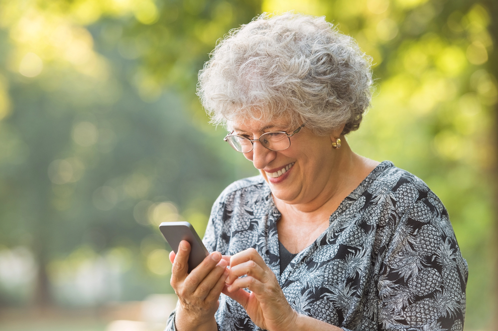 Smiling older woman with curly grey hair and glasses uses a mobile phone outdoors on a sunny day, surrounded by green trees in soft focus.