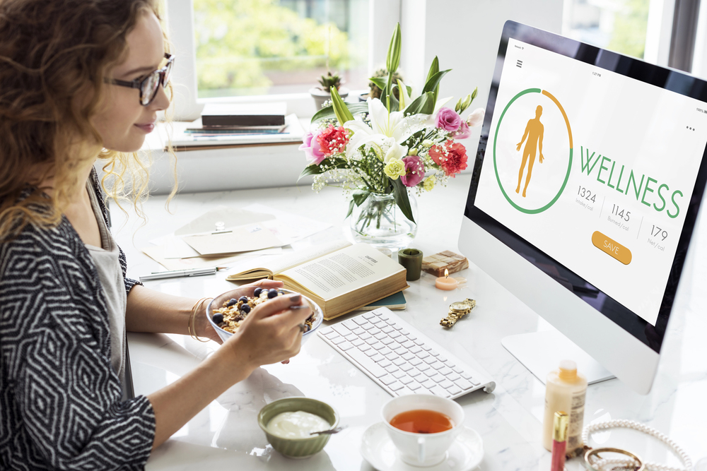 A woman with curly hair sits at a desk eating breakfast whilst looking at a wellbeing programme on a desktop computer. The desk has flowers, a keyboard, books, food, and a cup, creating a bright, organised workspace.