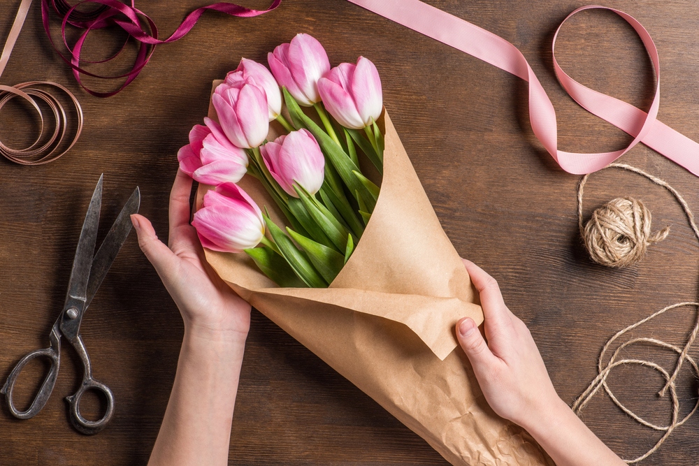 A person wraps a bouquet of pink and white tulips in brown paper on a wooden table, surrounded by scissors, string, and pink ribbons.