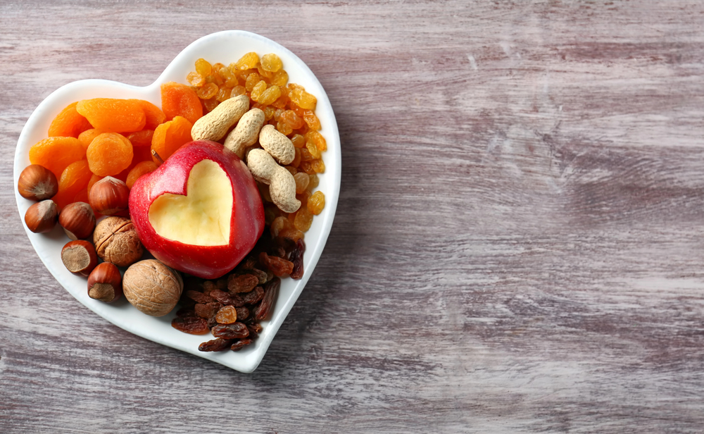 A heart-shaped dish filled with dried apricots, golden sultanas, peanuts, hazelnuts, walnuts, and a red apple with a heart-shaped cut-out, placed on a wooden surface.