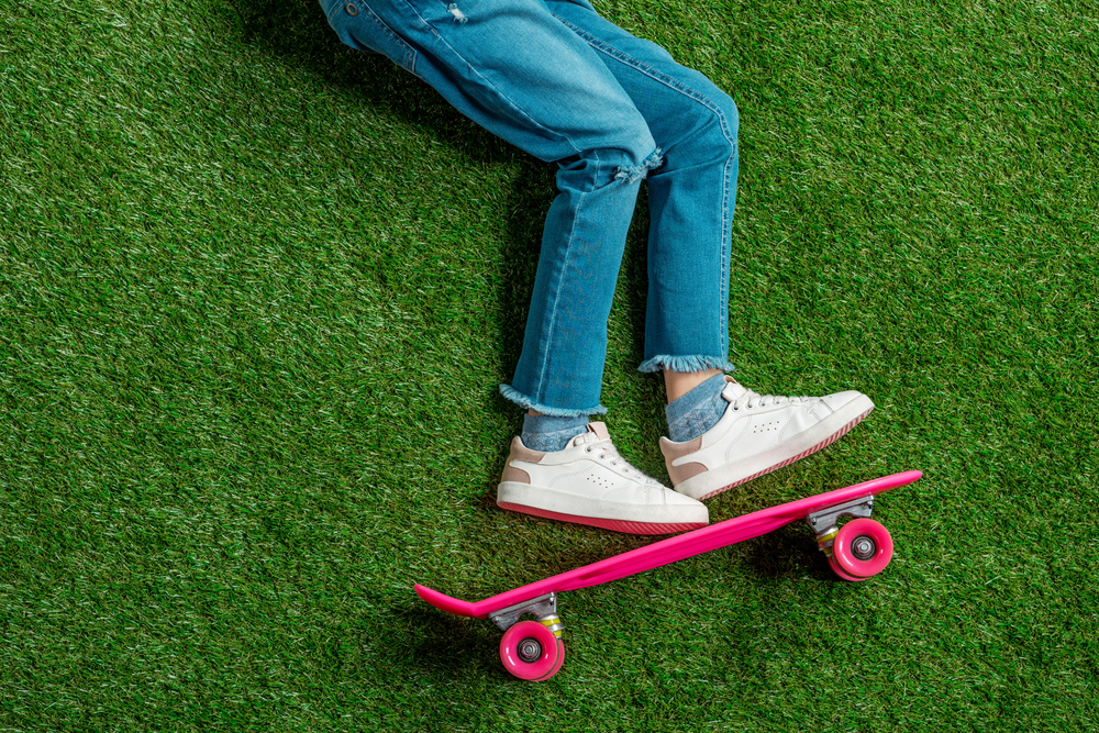 A person wearing blue jeans and white trainers rides a bright pink skateboard on green artificial grass, with only their legs visible in the image.