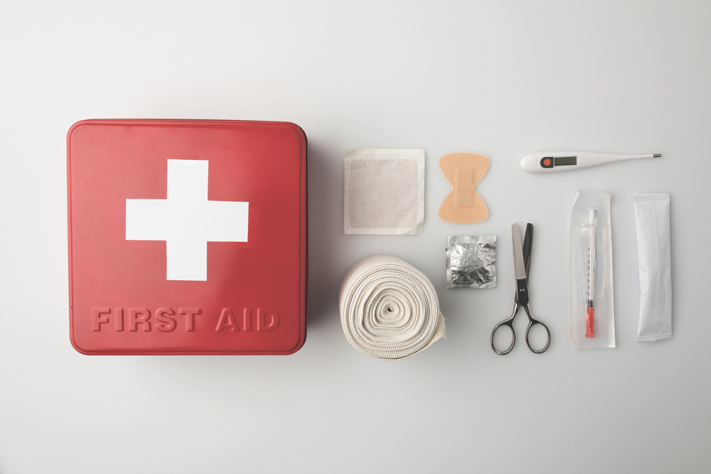 A red first aid kit with a white cross, bandage, adhesive plasters, gauze, a thermometer, medical scissors, a syringe, and a wrapped item, all neatly arranged on a white surface.