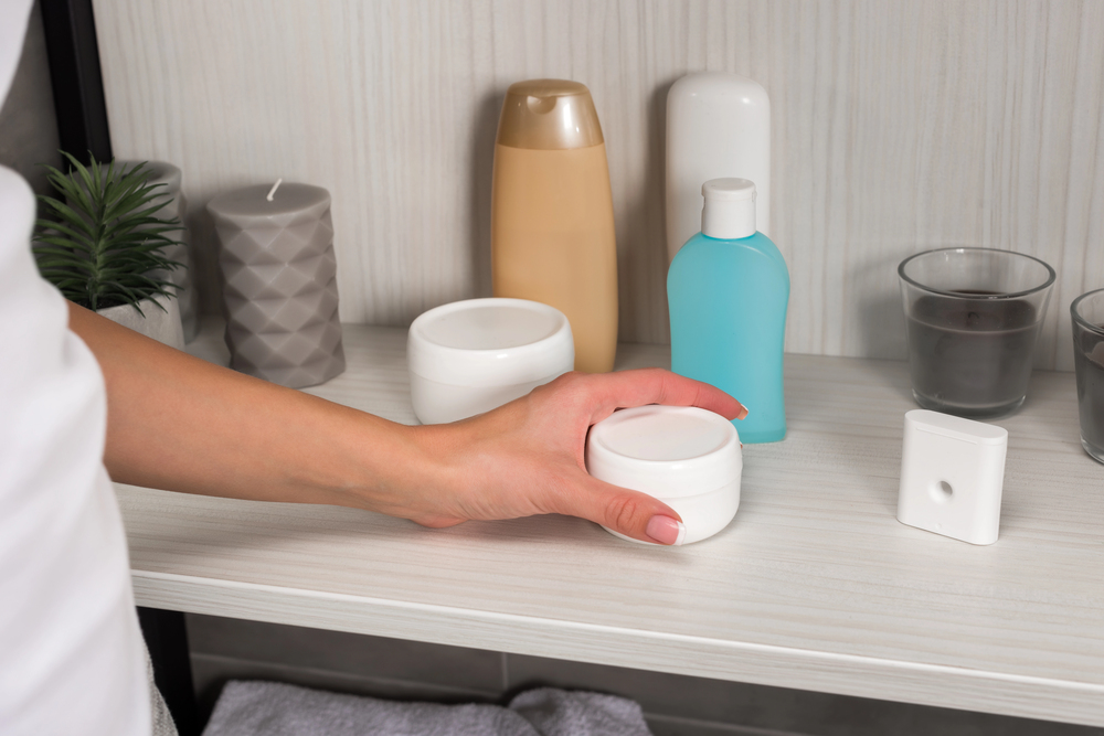 A person’s hand reaches for a small white jar on a bathroom shelf, which also holds bottles, containers, a candle, a plant, and two tumblers.