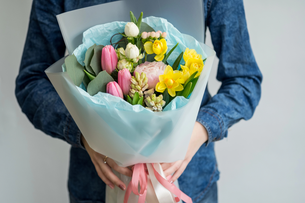 A person wearing a blue denim shirt holds a bouquet of colourful spring flowers, including pink tulips, yellow daffodils, and white blooms, wrapped in light blue paper and tied with a pink ribbon.