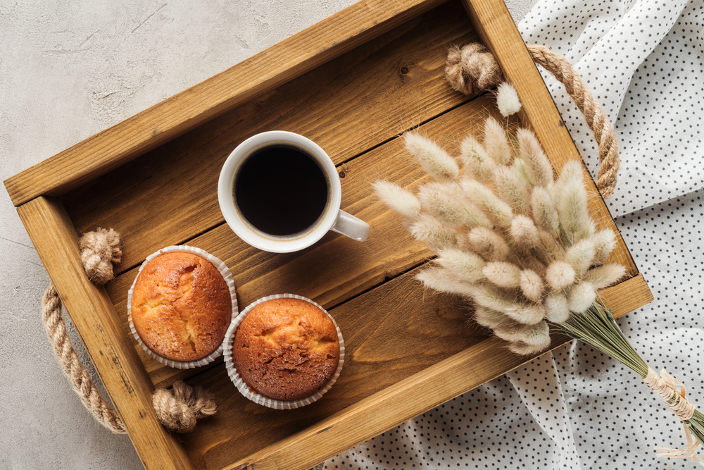 A wooden tray with a cup of black coffee, two muffins, and a bunch of dried light-coloured flowers, placed on a dotted fabric surface.