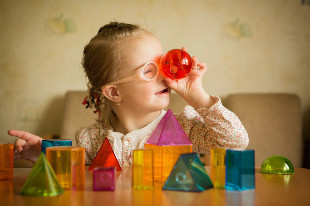 A young girl wearing glasses looks through a red transparent toy while sitting at a table with colourful geometric blocks, smiling and playing indoors.