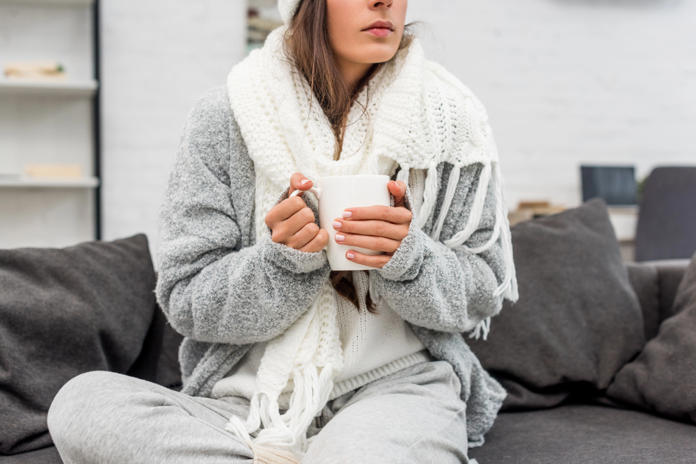 A woman wearing a cosy grey jumper, white scarf, and knitted hat sits cross-legged on a sofa, holding a white mug with both hands. The setting appears warm and comfortable.