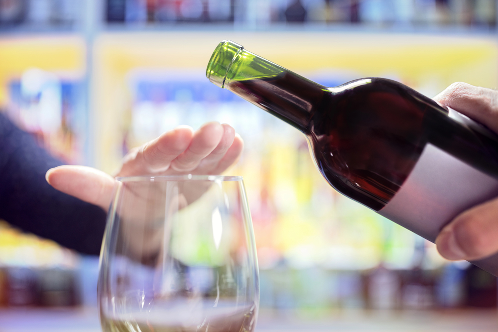 A person holds a wine bottle, about to pour into a glass, while another hand is raised with the palm facing down, signalling to stop or decline the wine. Shelves with bottles are blurred in the background.