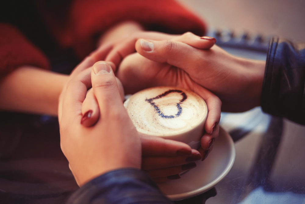 Two people hold hands around a cup of cappuccino with a heart shape drawn in cocoa powder on the foam, creating a warm and cosy atmosphere.