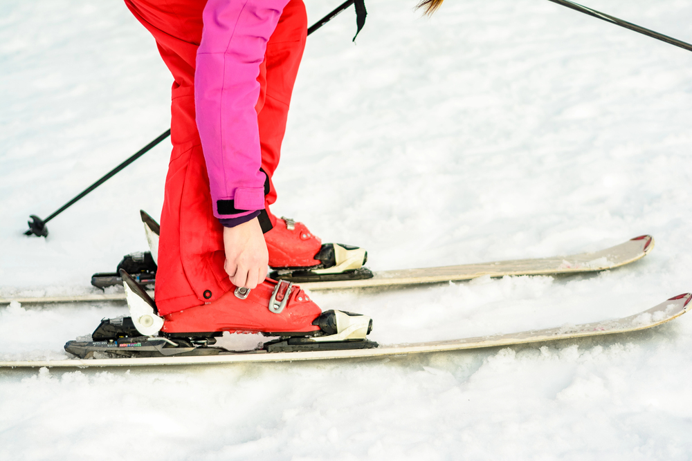 A person in red ski trousers and jacket adjusts their red ski boot on a pair of skis in the snow, holding a ski pole in one hand.