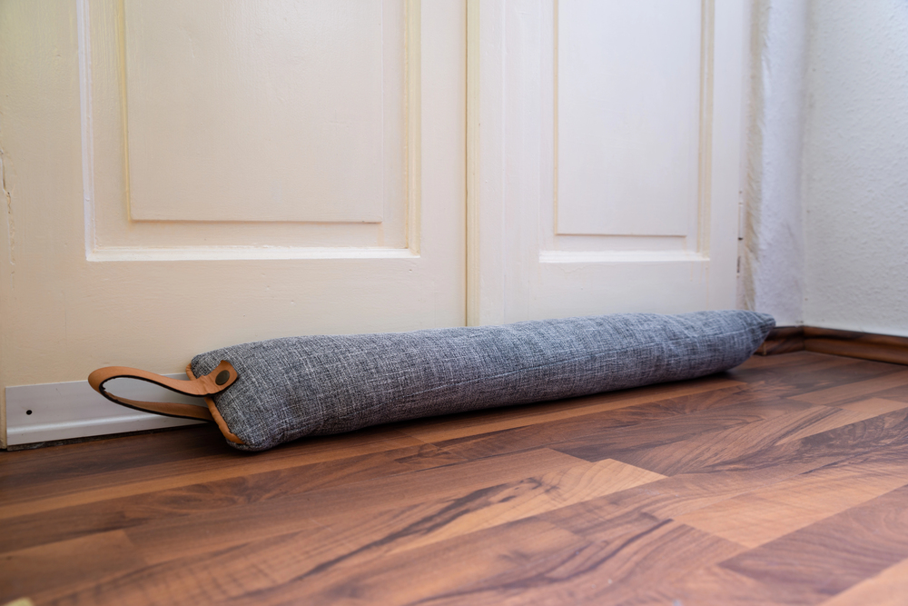 A grey fabric draught stopper with a leather handle lies along the bottom of a closed white door on a wooden floor, blocking the gap to prevent draughts.