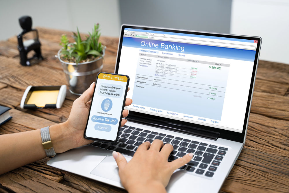 A person uses a mobile phone to approve a bank transfer while checking their online banking account on a laptop at a wooden desk. The laptop screen displays account balances and transactions.