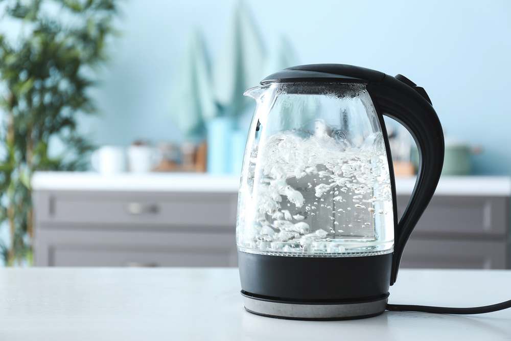 A glass electric kettle boiling water sits on a white kitchen worktop, with bubbles rising inside and a blurred kitchen background.