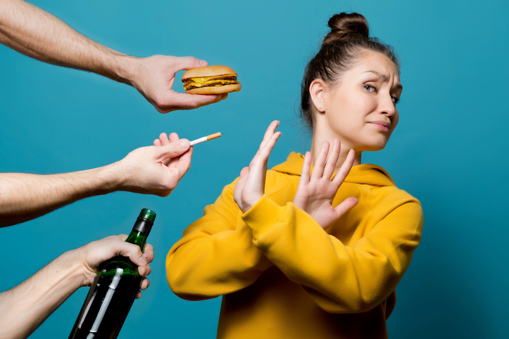 A woman in a yellow hoodie raises her hands to refuse a hamburger, cigarette, and bottle of alcohol being offered by three different hands against a blue background.