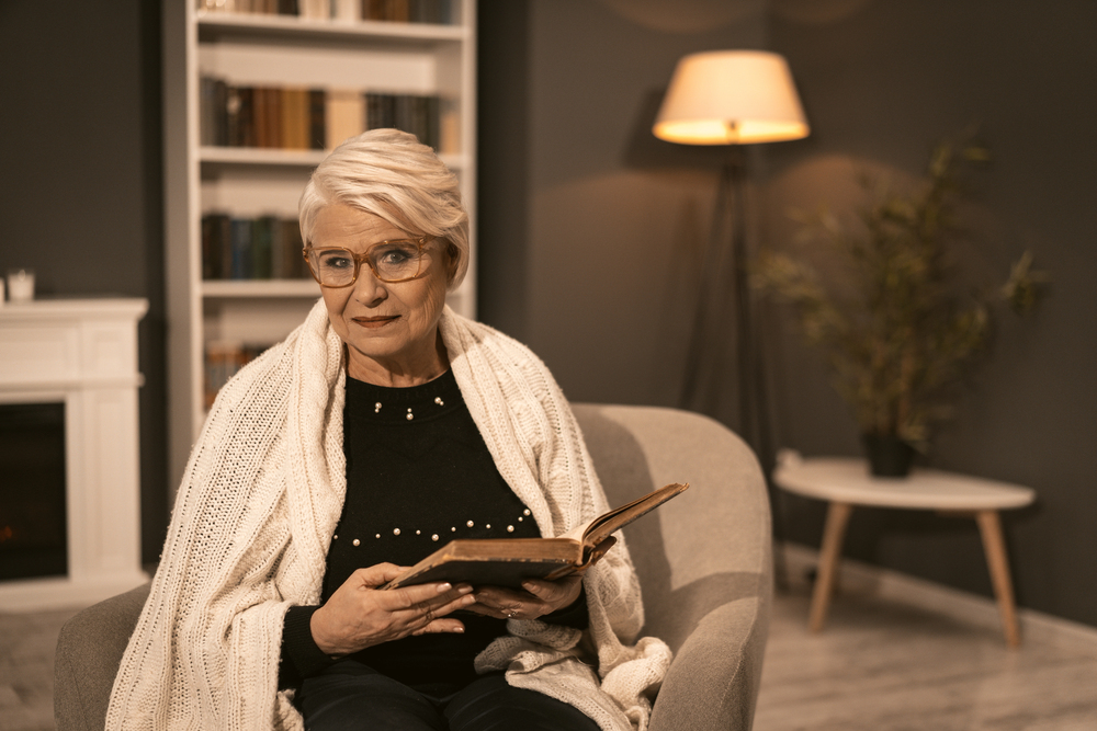 An elderly woman with short grey hair, glasses, and a white knitted shawl sits in an armchair holding an open book. She is indoors, with warm lighting, a bookcase, and a lamp in the background.