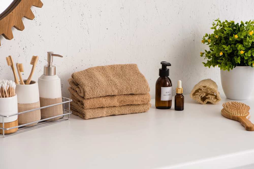 A bathroom worktop with folded beige towels, bottles of skincare products, a plant in a white pot, a wooden hairbrush, and a tray holding a soap dispenser, toothbrushes, and cotton buds.