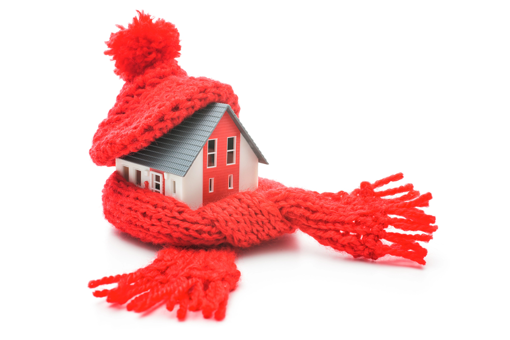 A small model house is wrapped in a red scarf and topped with a matching red knitted hat, symbolising warmth and protection, on a white background.